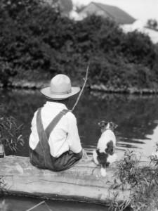 1920s 1930s Farm Boy Wearing Straw Hat And Overalls Sitting On Log With Spotted Dog Fishing